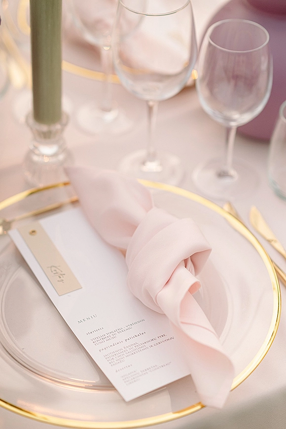 Wedding place setting with a blush napkin knot on a gold-rim plate and clear charger, menu and place card, wine glasses, taper candle on white linen