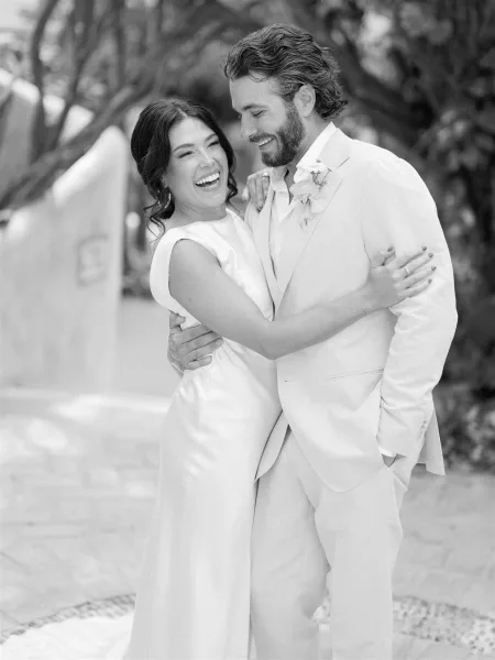 Couple portrait of a laughing bride and groom in close embrace, her minimalist gown and hoop earrings against trees by a stucco wall