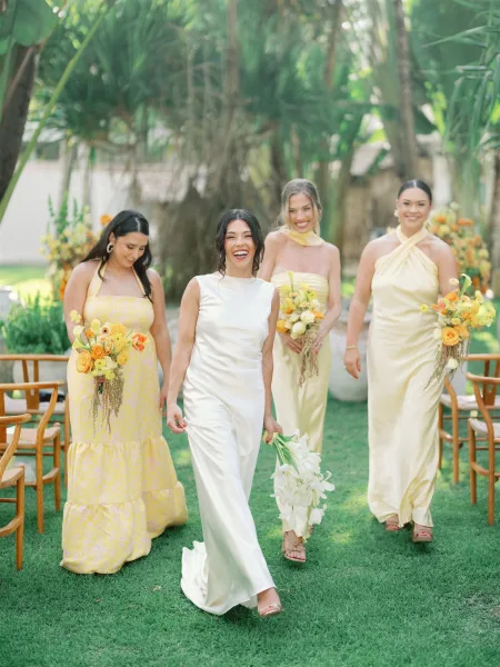Bridesmaid group photo with bridesmaids walking down aisle in mismatched yellow satin dresses, holding bouquets, palm trees behind