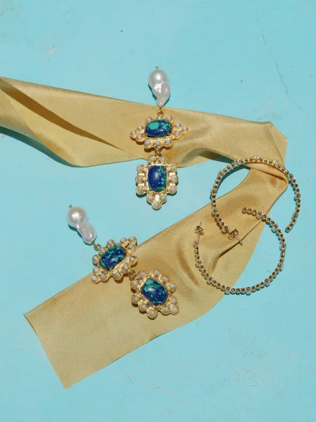 Wedding earrings with pearl drops and blue gemstone settings in gold, styled with champagne satin ribbon on a blue surface