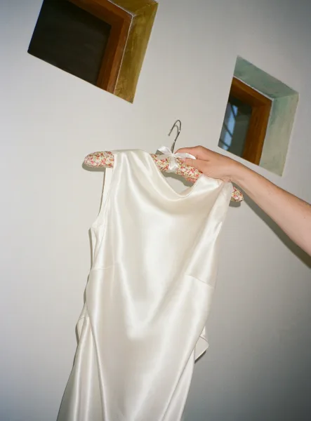 Wedding dress in satin sheen on a floral hanger with a white ribbon bow, held near windows against a white wall with wooden frames