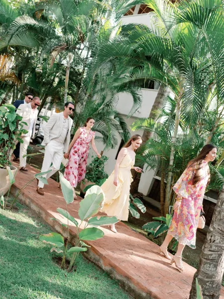Wedding guest style with tropical wedding guests walking a brick path past palm trees, wearing floral maxi dresses, linen suit, and sunglasses