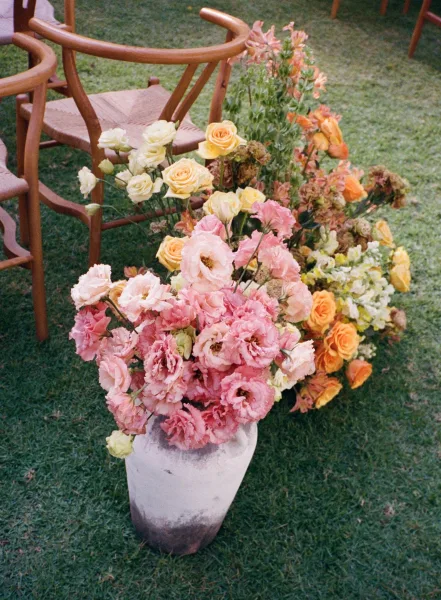 Ceremony aisle flowers in a ceramic vase beside wooden chairs, featuring pink lisianthus, roses, and hydrangea on a grass lawn