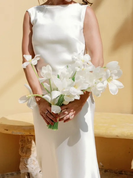 Bridal portrait of bride holding bouquet, a white flower bouquet and wedding rings against a sunlit stucco wall with shadows