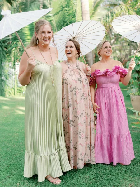 Bridesmaid group portrait of bridesmaids with parasols, laughing in colorful dresses on a lawn with tropical palm greenery behind