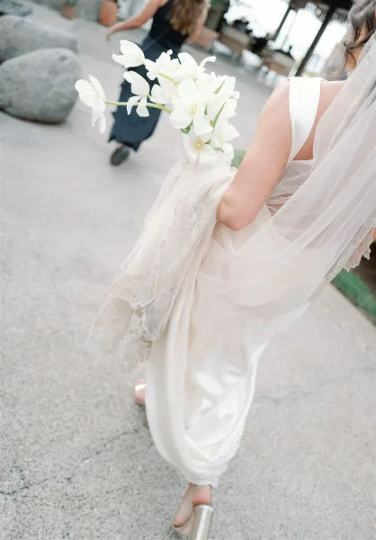 Bride walking from behind holding her wedding dress train, long veil trailing as she carries a white flower bouquet on a modern outdoor walkway