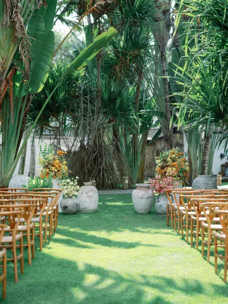 Ceremony setup with wood chairs lining a grass aisle, framed by large ceramic urn florals against tropical palm trees and lush greenery