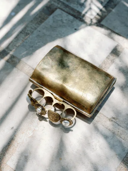Bridal jewelry flat lay featuring gold and pearl bracelets beside a vintage metal clutch on marble tile floor with sunlit shadows