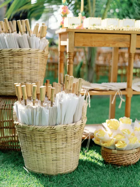 Wedding favor display with paper parasol favors in woven baskets on a wooden table, scattered petals and cloth runners in a tropical garden lawn