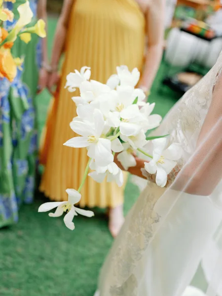 Bridal bouquet of white flowers held by a bride in lace wedding dress and veil, with green lawn and wedding guests behind