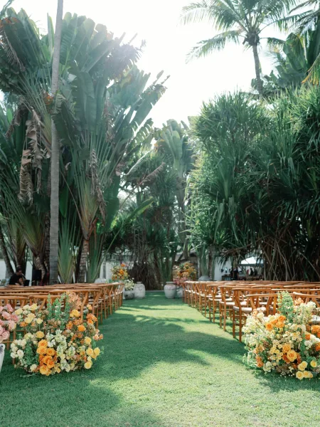 Outdoor ceremony setup with tropical wedding ceremony seating, wood chairs lining a grass aisle with rose florals in ceramic urns and palms beyond