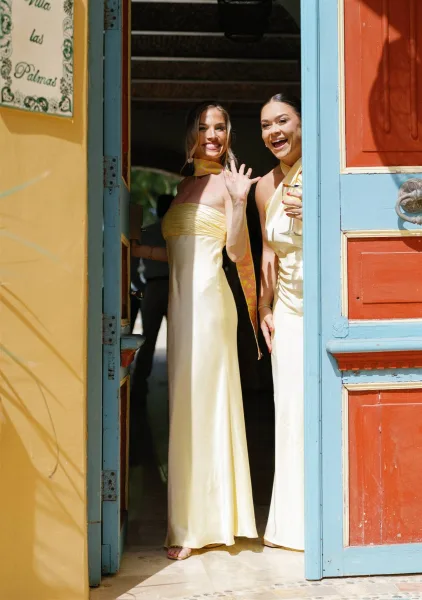 Bridesmaid portrait of two women in satin dresses holding a champagne flute by a colorful doorway, one with a neck scarf in soft shadows