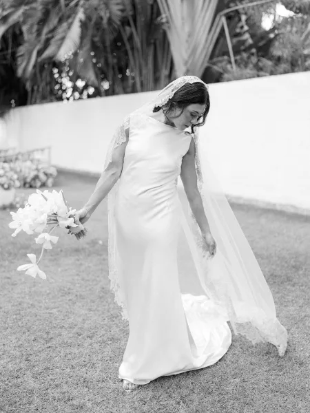 Bridal portrait of a bride in a simple satin wedding dress with lace veil, holding a bouquet on a grassy lawn by palm trees