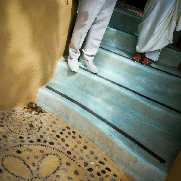 Wedding shoes beside bride and groom shoes, white loafers and bridal heels framed by dress hem on blue stone steps near a doorway shadow