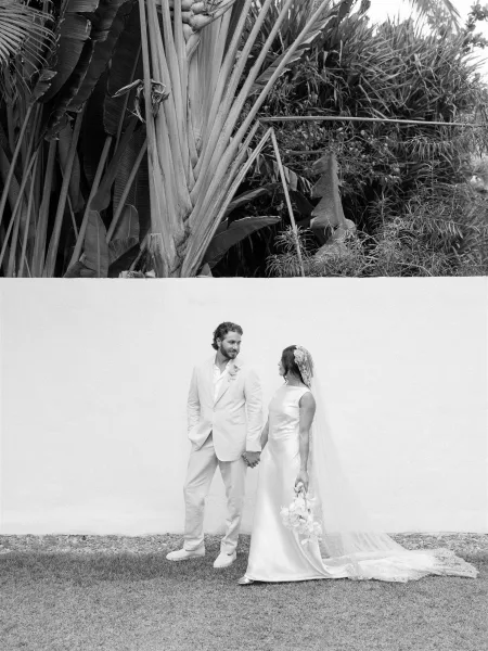 Couple portrait of bride and groom holding hands, her lace-edged veil and orchid bouquet flowing beside a white wall with tropical foliage