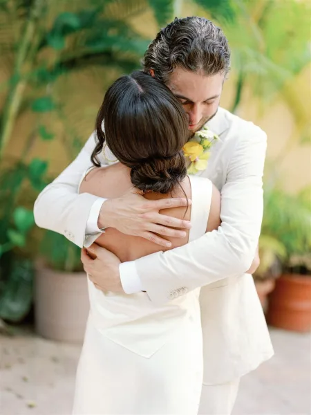 Wedding couple embrace in a first look hug, groom in a white suit with yellow boutonniere holding bride in sleeveless dress on greenery walkway