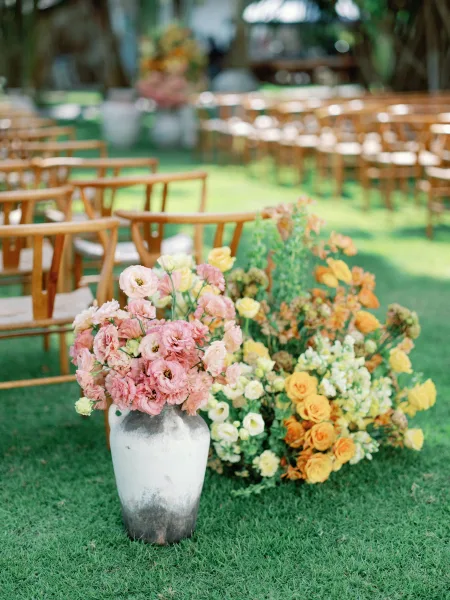 Ceremony aisle decor with outdoor ceremony seating of wood chairs and pastel floral arrangements in ceramic vases on a garden lawn under trees