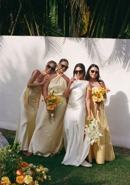 Bridesmaids portrait with bride, in mix-and-match yellow dresses holding bouquets and sunglasses by a white stucco wall with palms