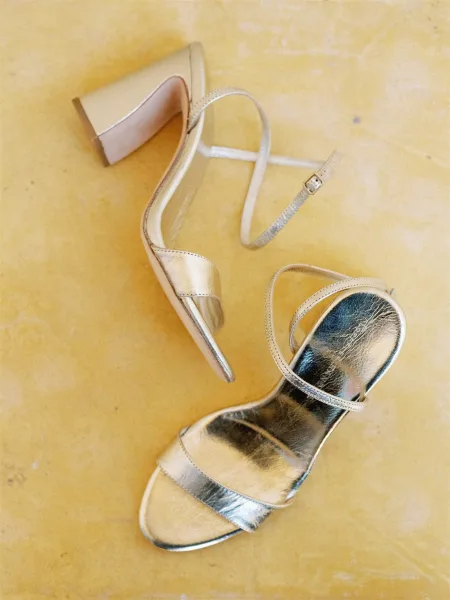 Wedding shoes, bridal heels with metallic strappy design, ankle straps, and buckles on a yellow textured studio floor