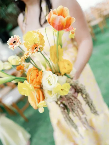 Bridal bouquet of orange and yellow bouquet blooms with tulips and greenery, paired with sunglasses on an outdoor lawn by a wooden chair