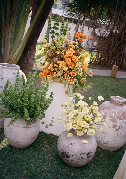 Wedding floral arrangements with ceremony pedestal flowers featuring orange, yellow, and white roses with greenery along a garden walkway outdoors