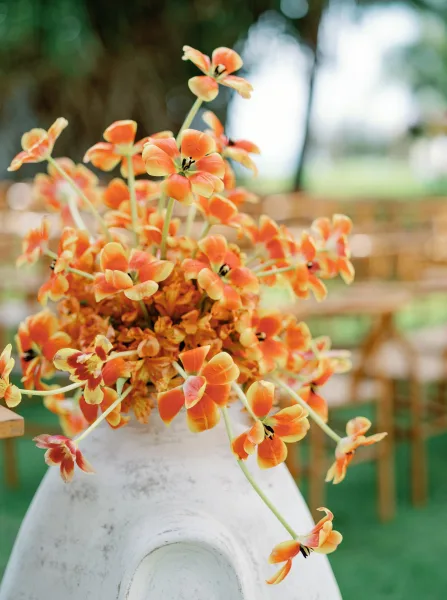 Wedding centerpiece with orange floral centerpiece blooms in a white ceramic vase, set before outdoor reception chairs on a green lawn under trees