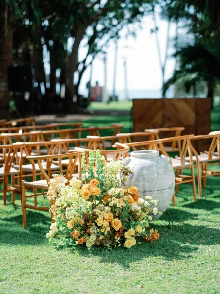 Ceremony setup with wooden chairs and floral arrangement in a large ceramic urn on a grass lawn, with palm trees and ocean view beyond