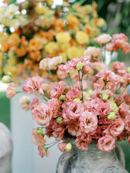 Wedding floral arrangement with pink wedding centerpiece blooms and green buds in a textured ceramic vase, set against blurred orange-yellow florals