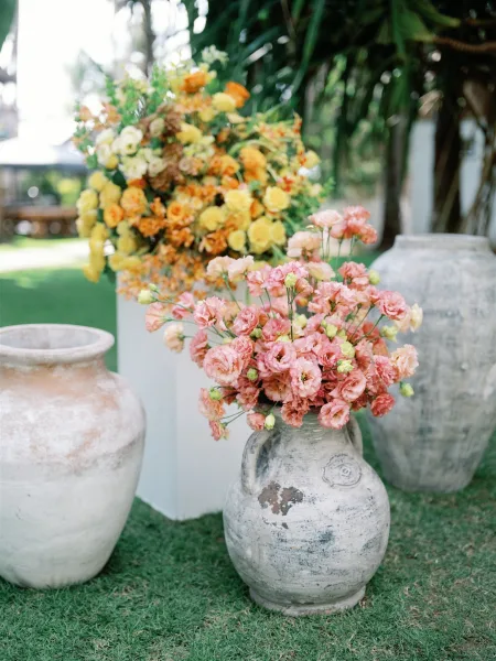 Wedding floral arrangements with ceremony entrance florals in large urns of pink, yellow, and orange blooms on a garden lawn with palm leaves
