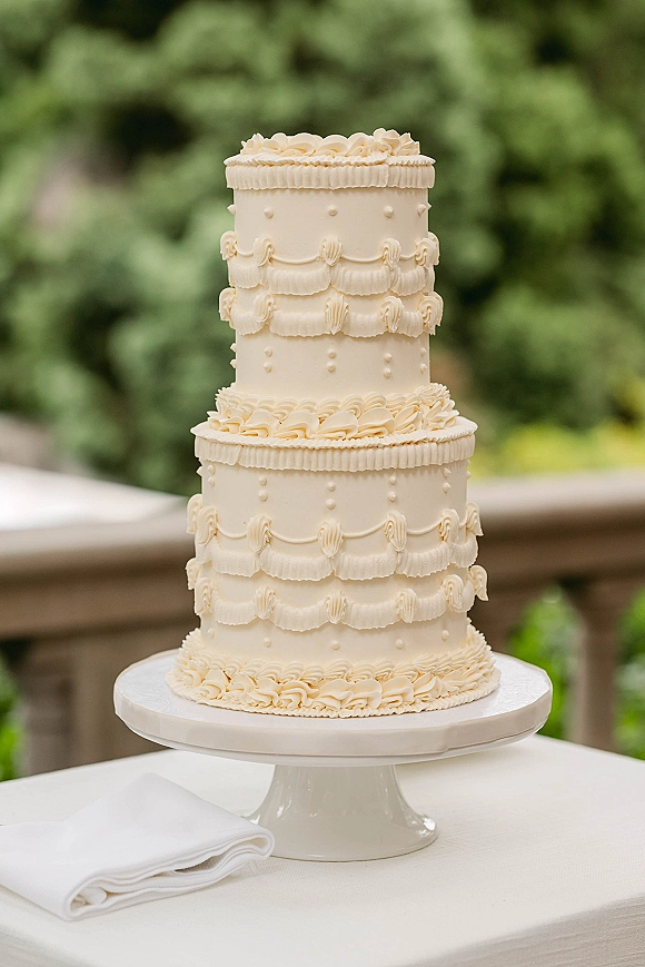 Wedding cake with three tier wedding cake design, ivory buttercream ruffles and scalloped piping on a stand, set against garden greenery