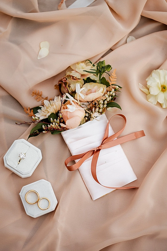 Wedding flat lay with wedding invitation, ribbon bow, peach roses, daisies, greenery, and rings on a blush fabric drape backdrop
