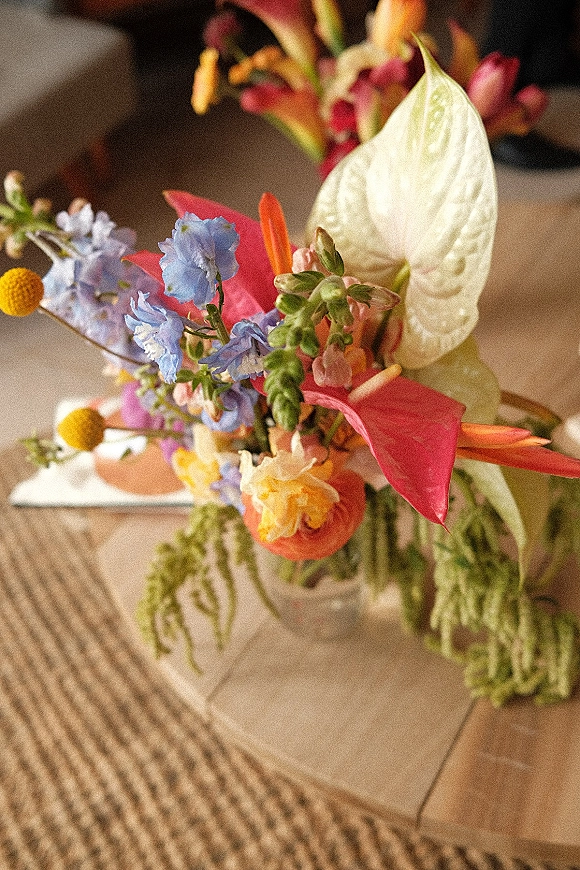 Wedding centerpiece with colorful florals in a clear glass vase, featuring anthurium, blue delphinium, daisies, and a white taper candle on tablecloth