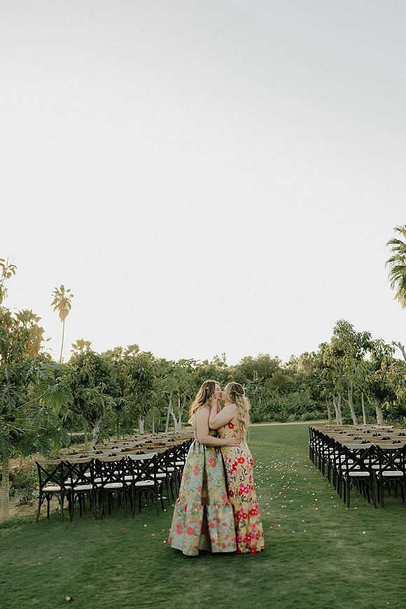Wedding kiss between a lesbian wedding couple in a chair-lined aisle with flower petals on a grass lawn framed by orchard and palm trees