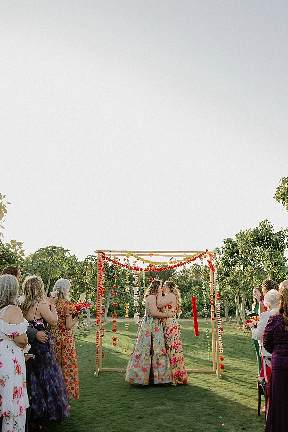 Ceremony kiss at a same sex wedding ceremony as two brides kiss beneath a wooden arch with hanging florals on a garden lawn
