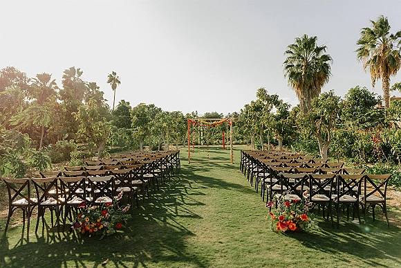 Outdoor ceremony setup with wood cross-back chairs lining an aisle runner, bright floral arrangements and garlanded arch on an orchard lawn