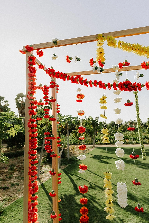 Floral mandap decor for an Indian wedding mandap with a wooden frame draped in hanging marigold and red-white flower garlands on a garden lawn