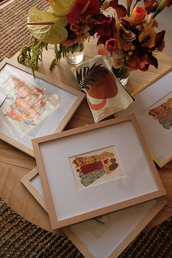 Wedding guest book display with framed artwork and watercolor illustrations beside colorful flowers in glass vases on a wood table over a woven rug