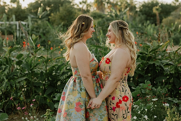 Couple portrait of two brides holding hands in matching floral print dresses, rings visible, framed by garden greenery, wildflowers, and palm trees
