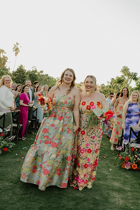Wedding recessional as two brides walk the outdoor aisle past cheering guests, holding bouquets with scattered petals and aisle florals