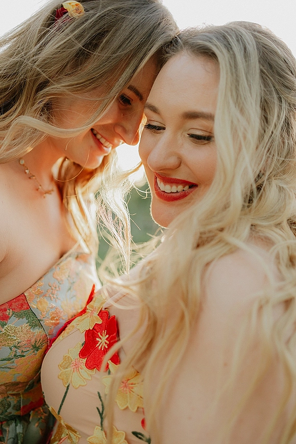 Couple portrait of two brides touching foreheads in a lesbian wedding portrait, one in a floral dress with red lipstick against bright greenery