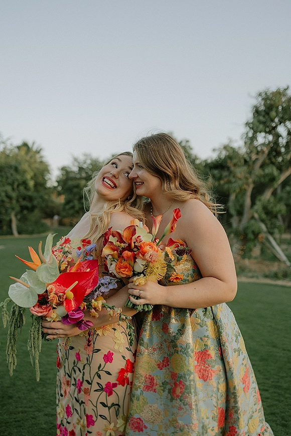 Bridesmaid portrait of women holding bouquets, with colorful flowers and floral dresses on a grass lawn with trees and open sky