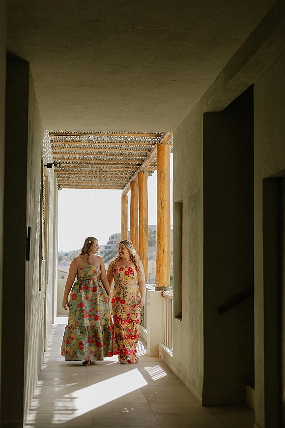 Couple walking portrait of two brides holding hands in colorful floral maxi dresses along a sunlit pergola corridor with wooden columns