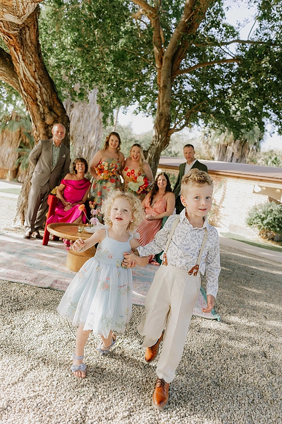 Flower girl and ring bearer holding hands, ring bearer suspenders outfit beside bridesmaids with bouquets under large trees on patio