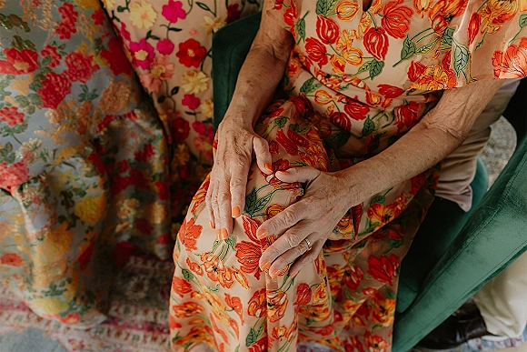Wedding guest outfit in an orange floral wedding guest dress, close-up of hand with engagement ring and orange nail polish on green velvet chair