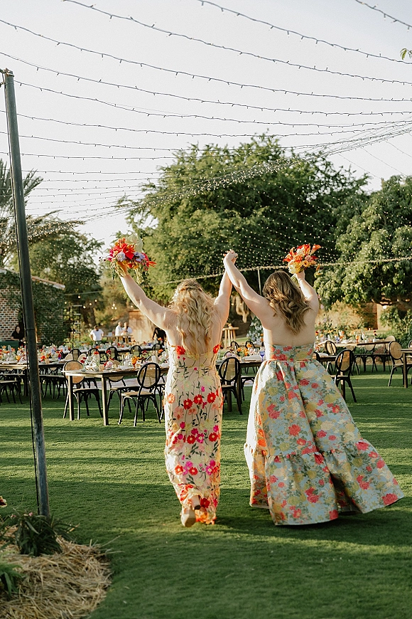 Wedding reception moment with guests holding hands and raised bouquets in floral dresses under string lights at outdoor lawn tables