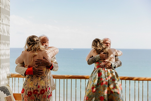 Family wedding hug as the bride embraces her father on a sunlit seaside terrace, guests in floral dresses and suits near a balcony railing