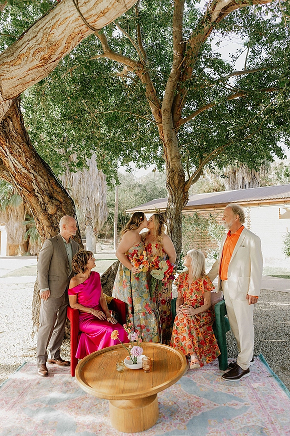 Couple portrait of two women kissing beside a cocktail lounge on a lawn, with a colorful wedding bouquet and brick building backdrop