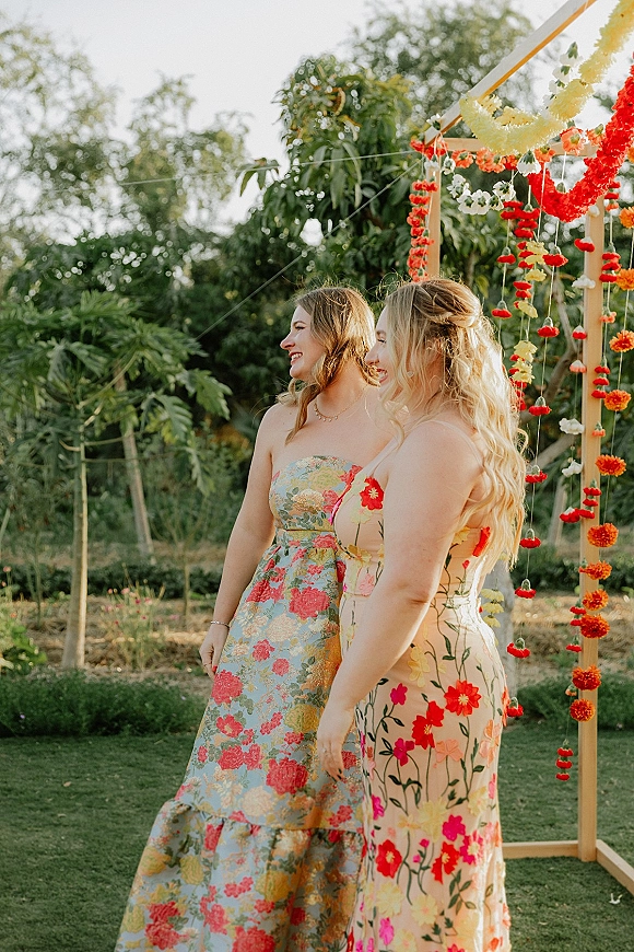 Wedding guest portraits of women in bridesmaid floral dresses under hanging flower garlands on a wooden frame in a garden lawn setting