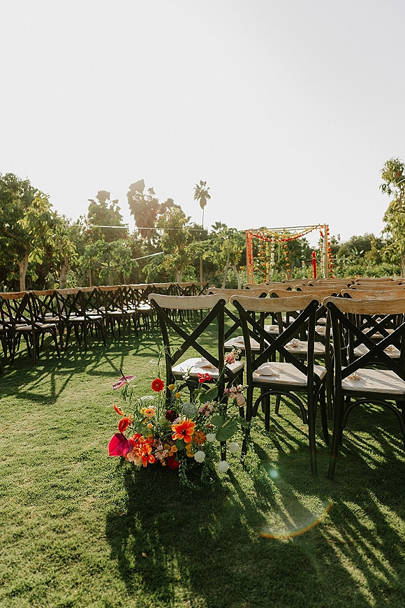 Outdoor ceremony setup with wedding ceremony chairs lining a grassy aisle, colorful florals, and a fabric-draped arch beneath trees in sunlight