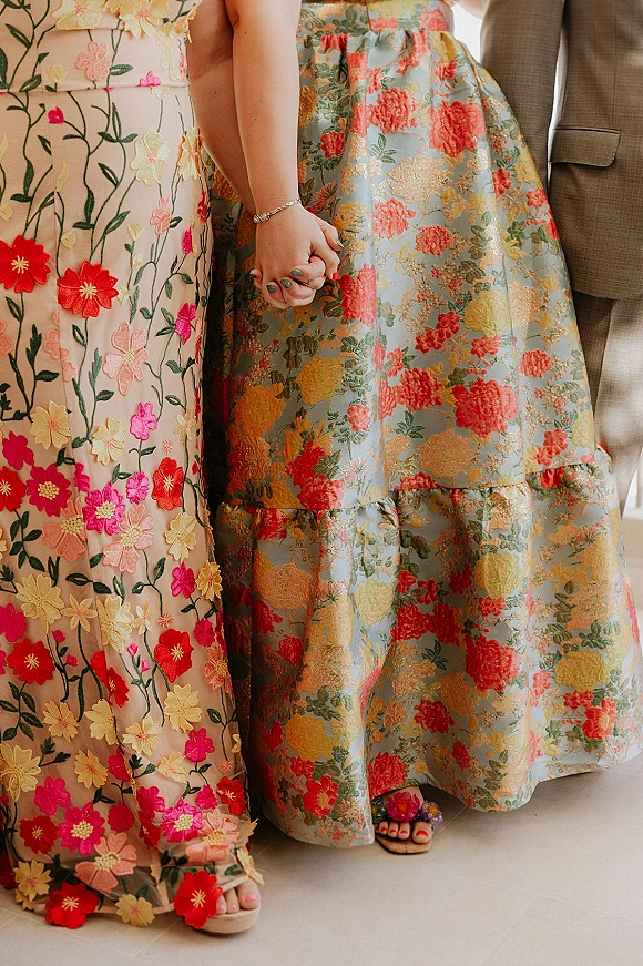 Holding hands photo of two women in floral wedding guest dresses, with bracelet and embroidered fabric details above an indoor floor